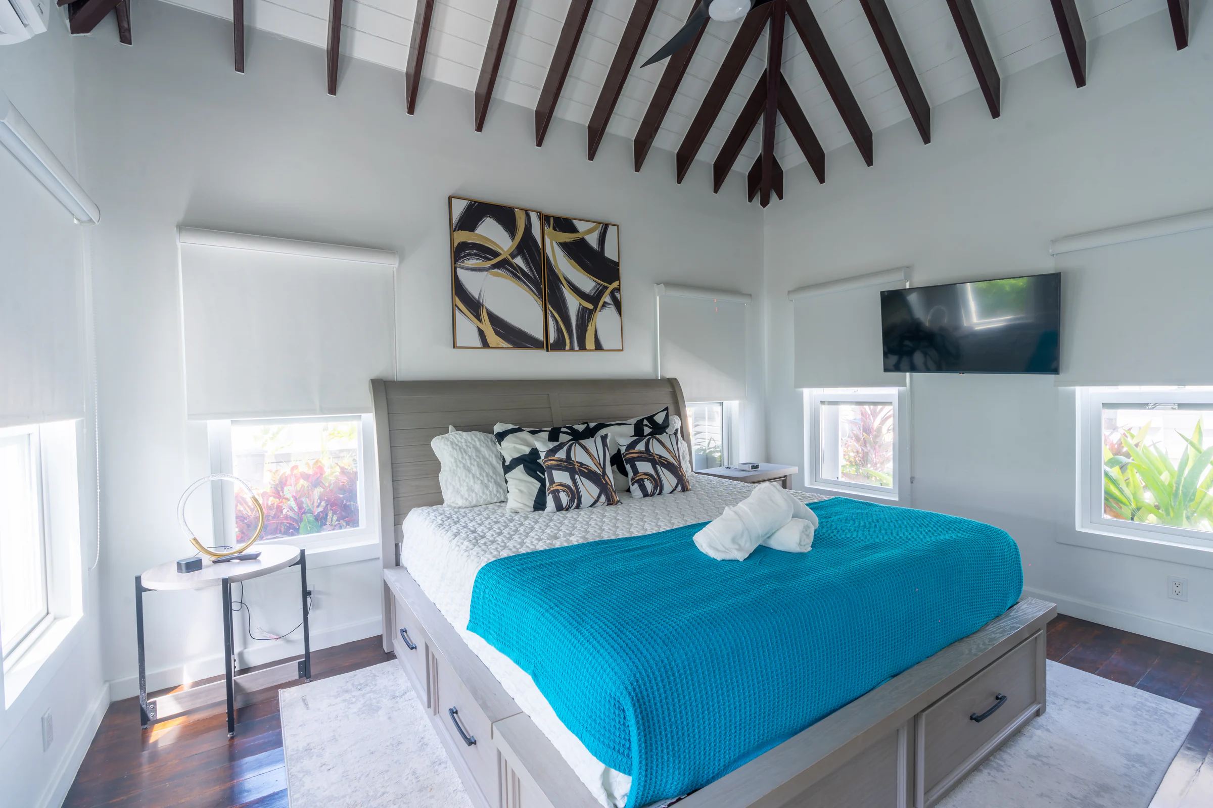 A king bed made up in white and turquoise beneath a dark timber ceiling fanning out from a central beam, windows on three sides looking into planting.