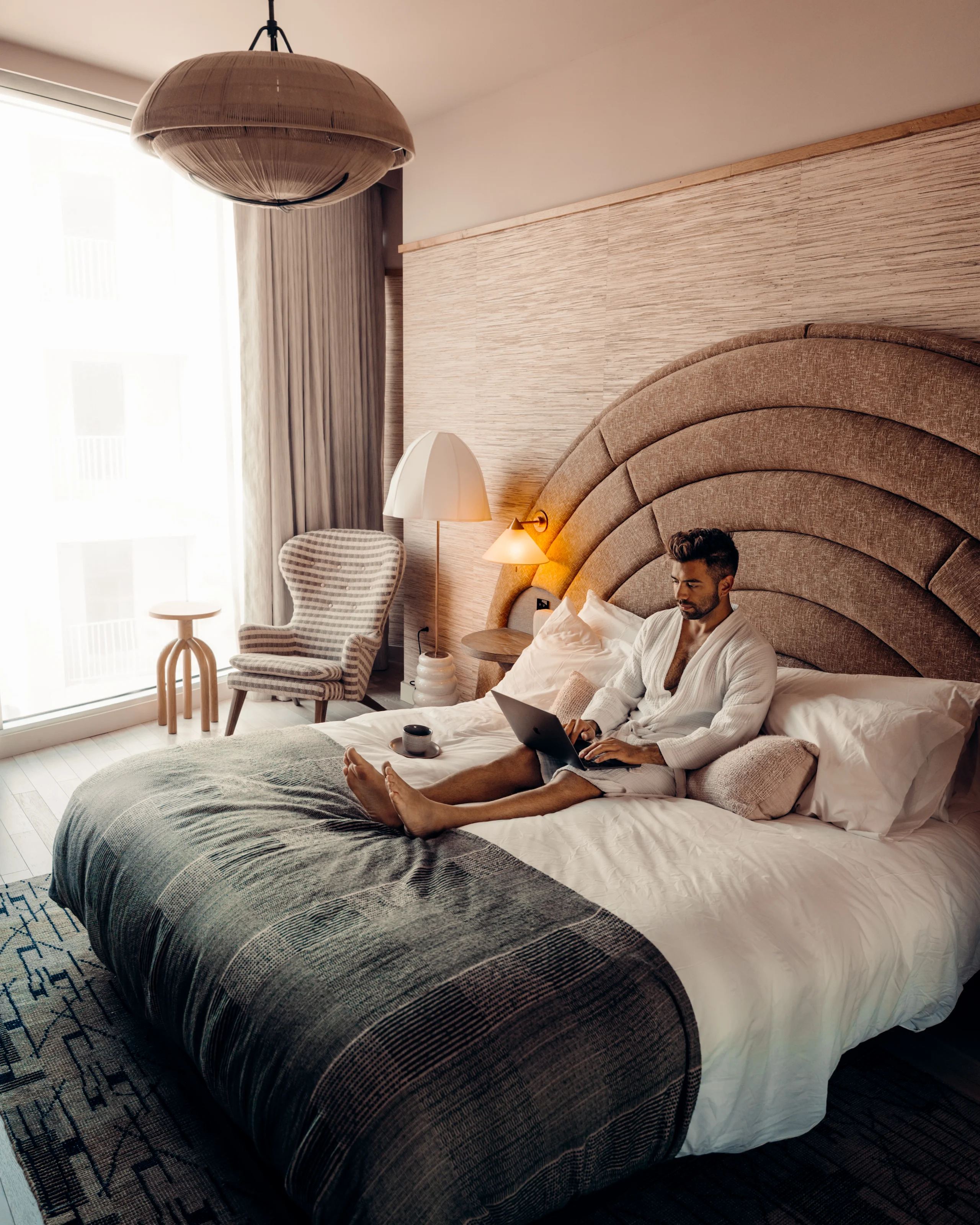A guest in a robe working on a laptop on the bed of a light hotel suite