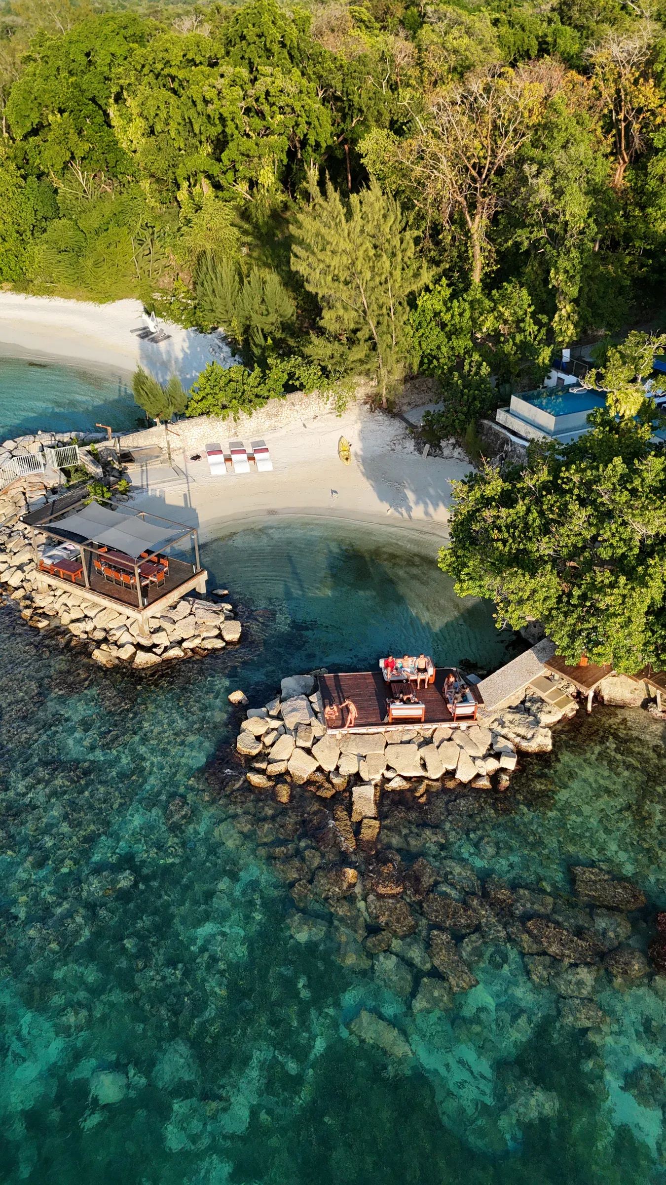 A rocky cove from the air: a shaded dining deck out on the rocks, a timber deck of loungers over the water, a small sand beach, and an infinity pool set back in the trees.