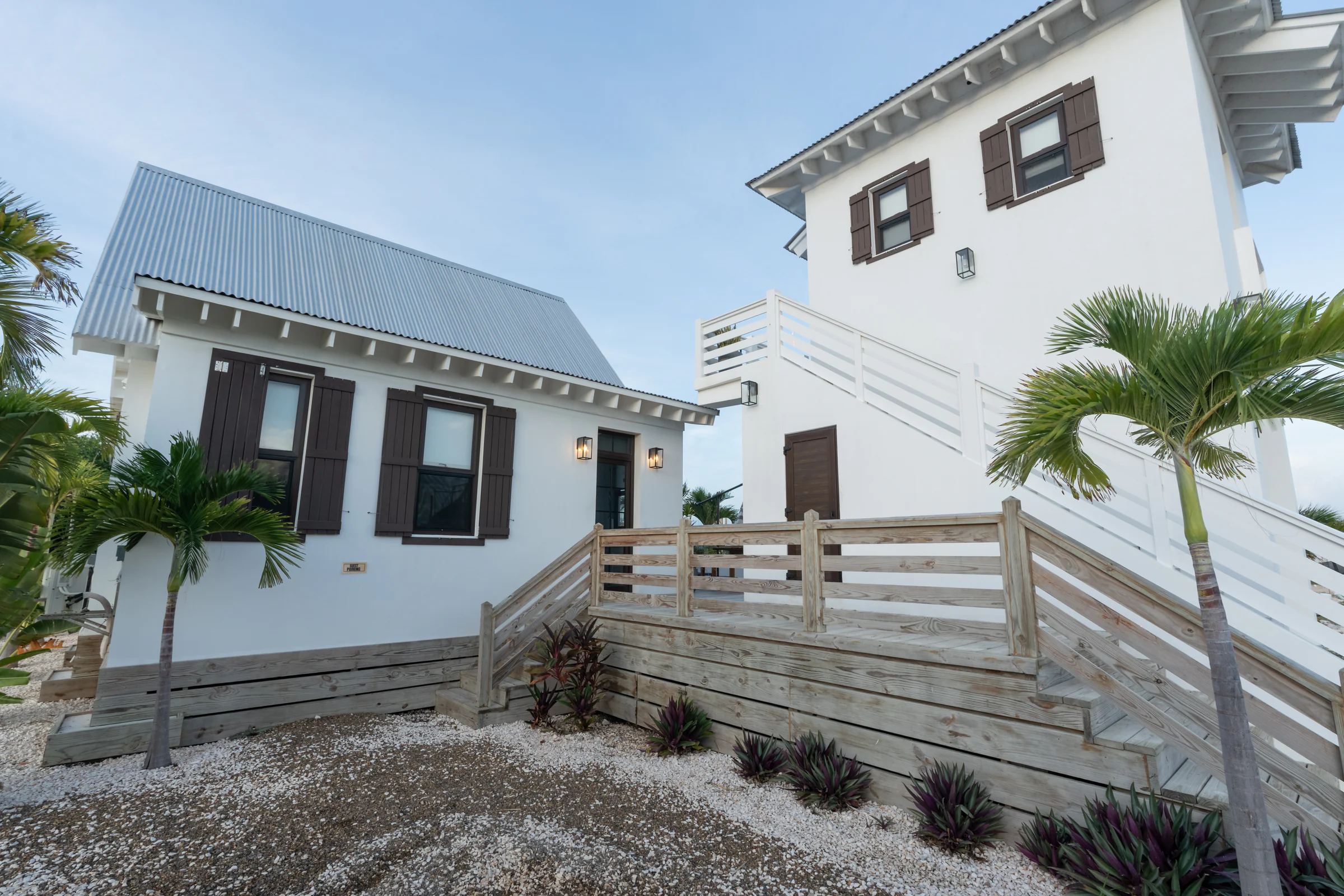 Two white cottages with dark shutters and metal roofs, joined by a weathered timber deck and steps, young palms in a bed of white gravel.