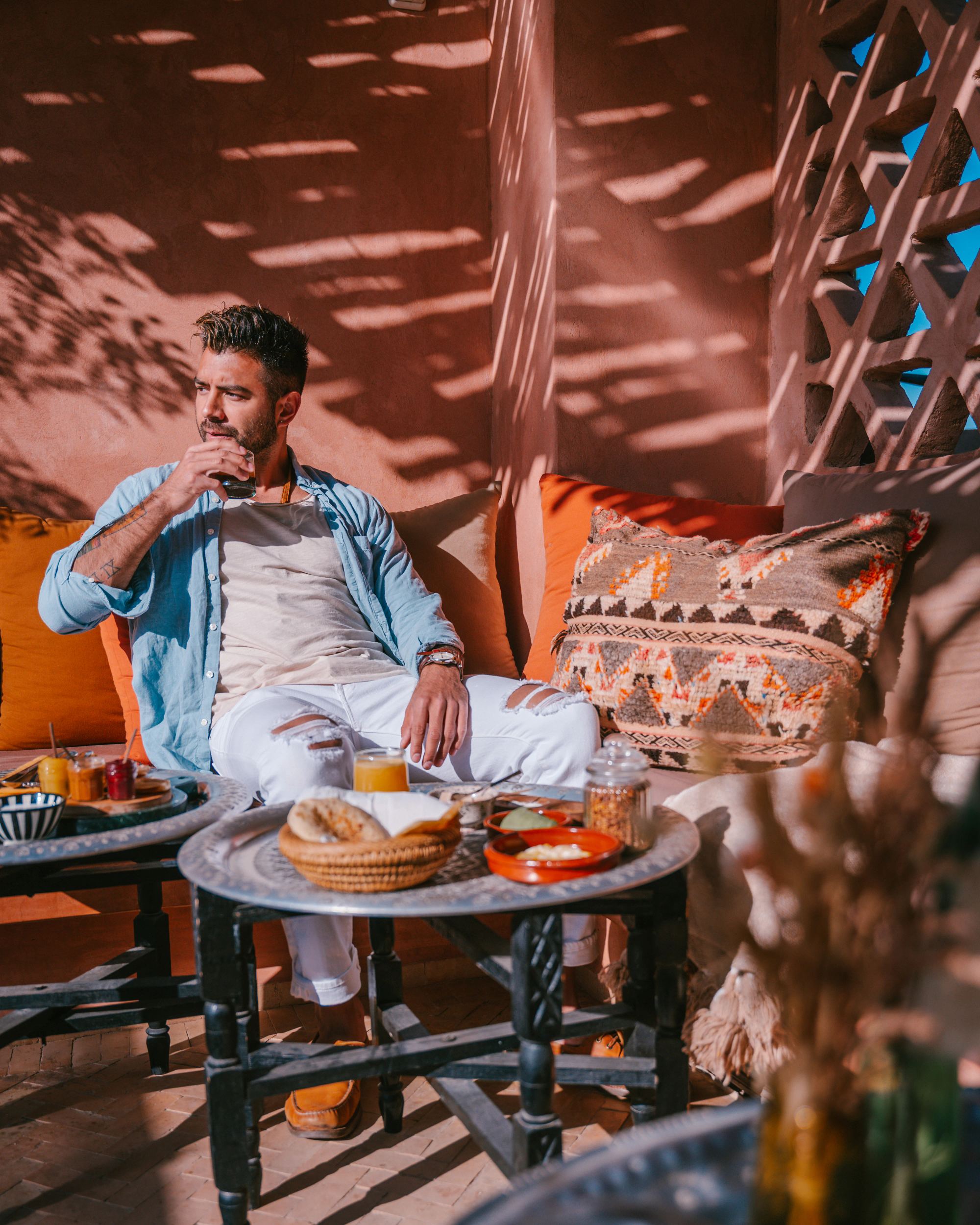A guest on a terracotta terrace with a drink, breakfast laid out on engraved tray tables, a pierced screen throwing shadows across the wall