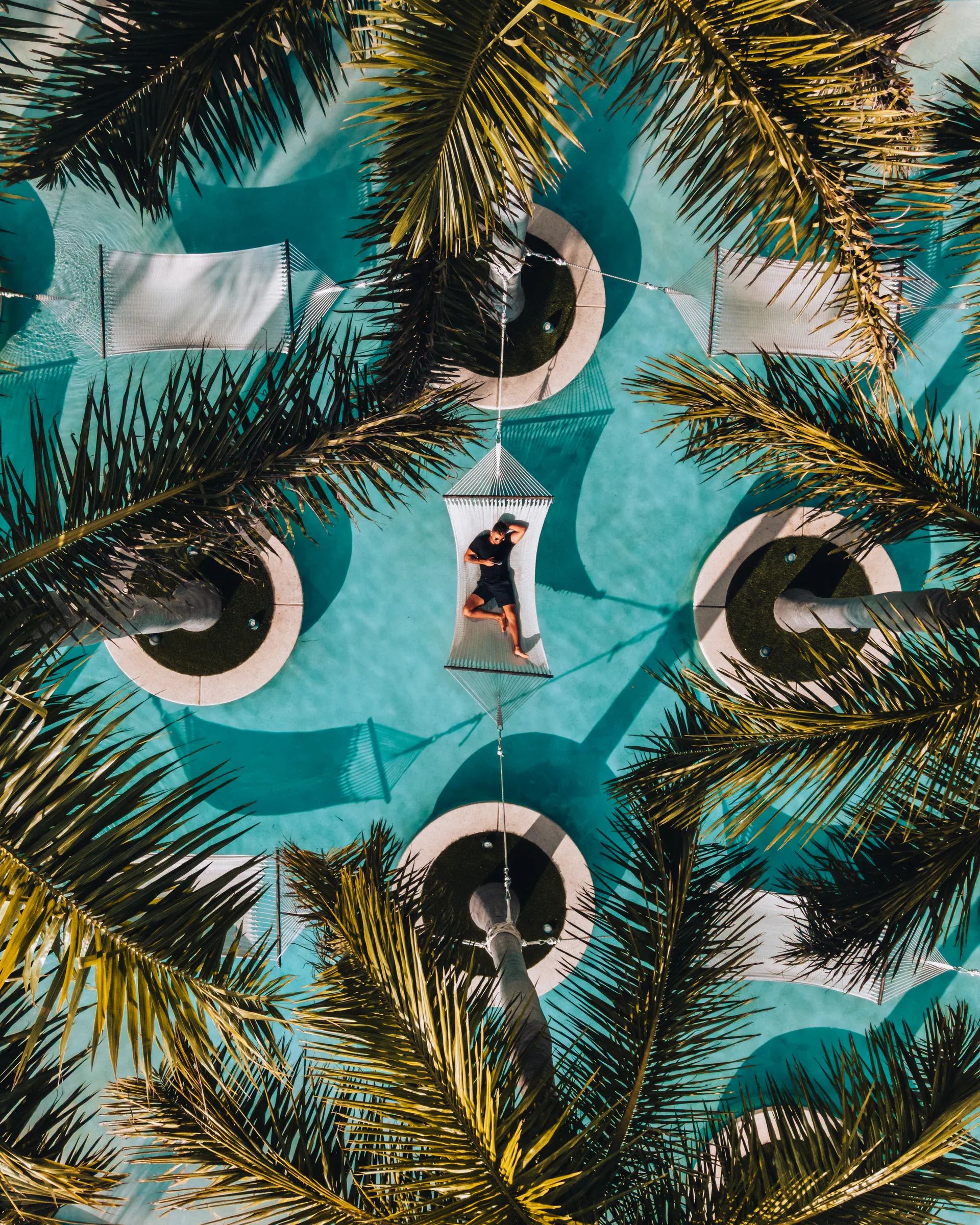 Straight down over a turquoise pool, white string hammocks slung between round planters of palms, a guest lying in the centre one, palm fronds filling the corners of the frame.