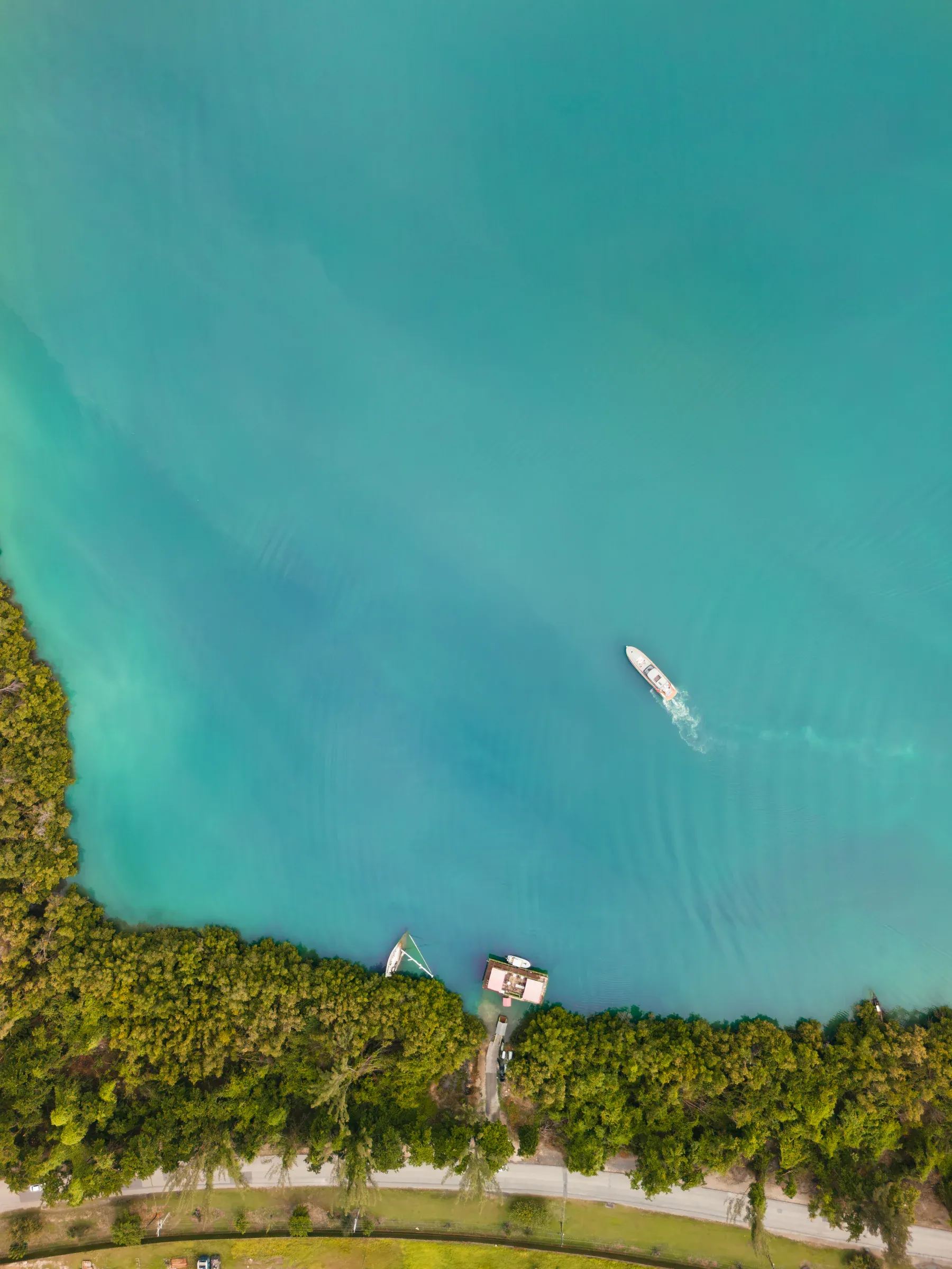 Straight down over shallow turquoise water, a small motorboat drawing a wake, a dock and a moored sailboat against the treeline.