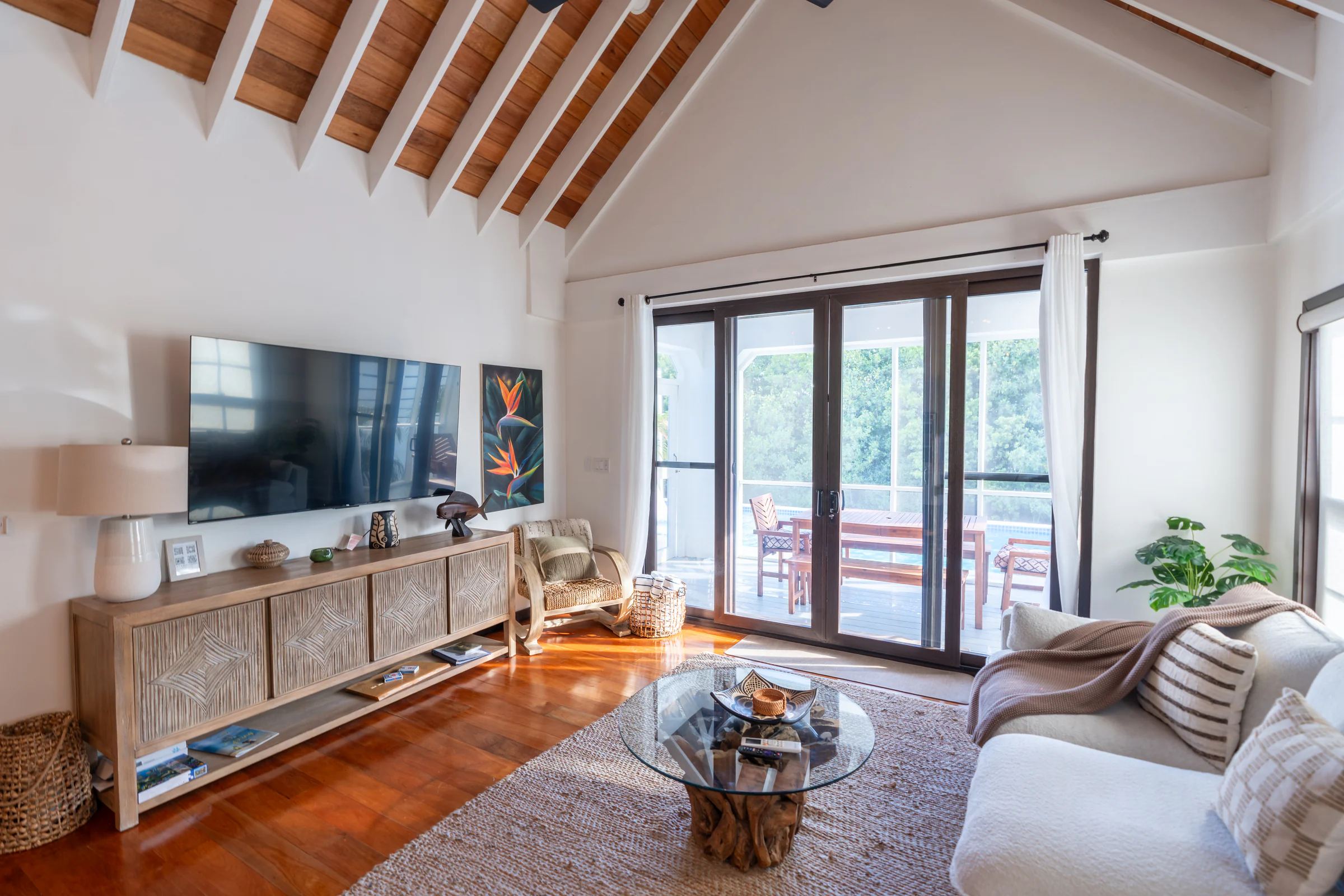 A living room under a vaulted ceiling of exposed timber, sliding doors open onto a deck and the pool beyond, a low carved cabinet and a glass table on a jute rug.