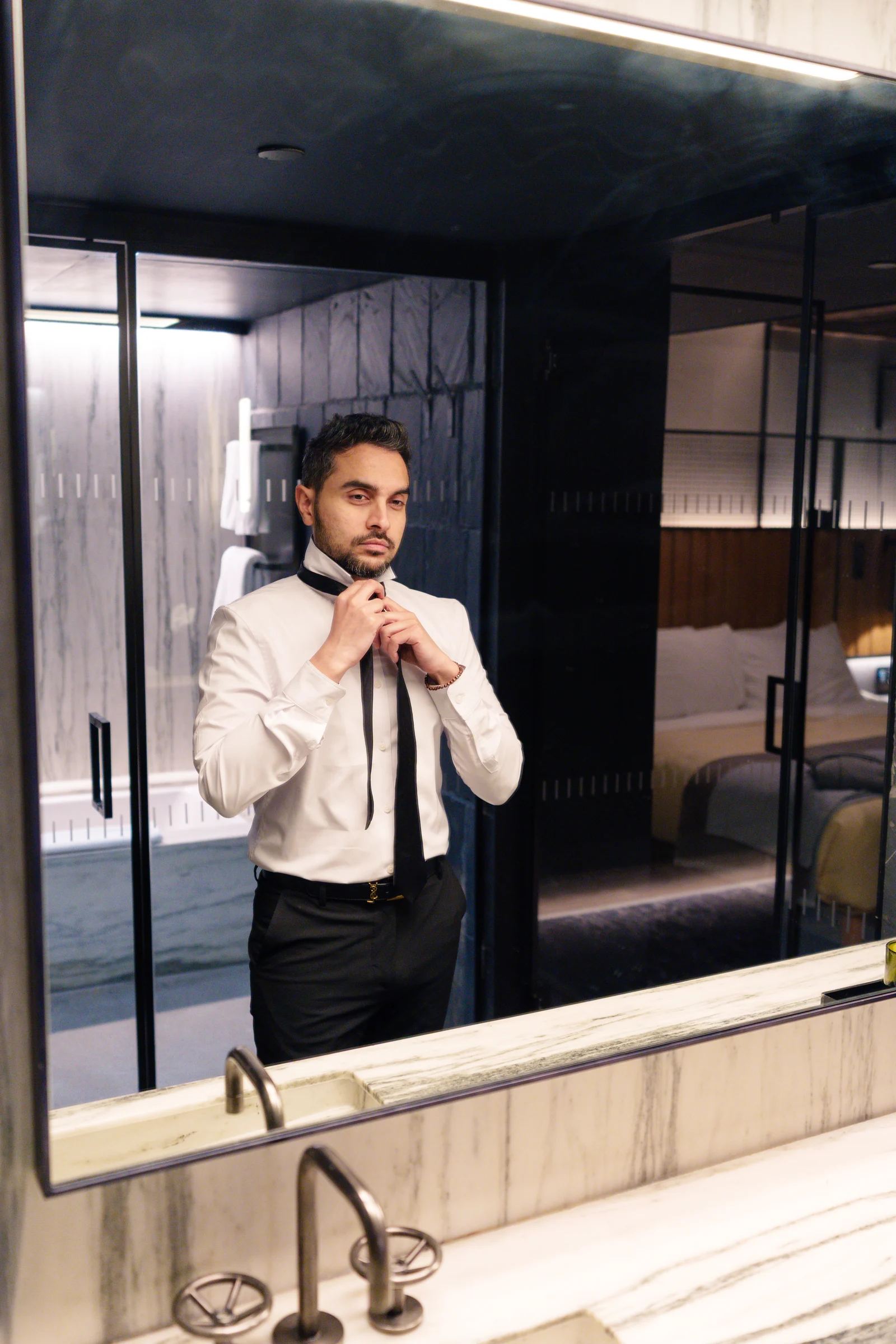 A guest tying a black tie, reflected in the lit mirror of a dark marble hotel bathroom, the bed and window of the room visible behind him.