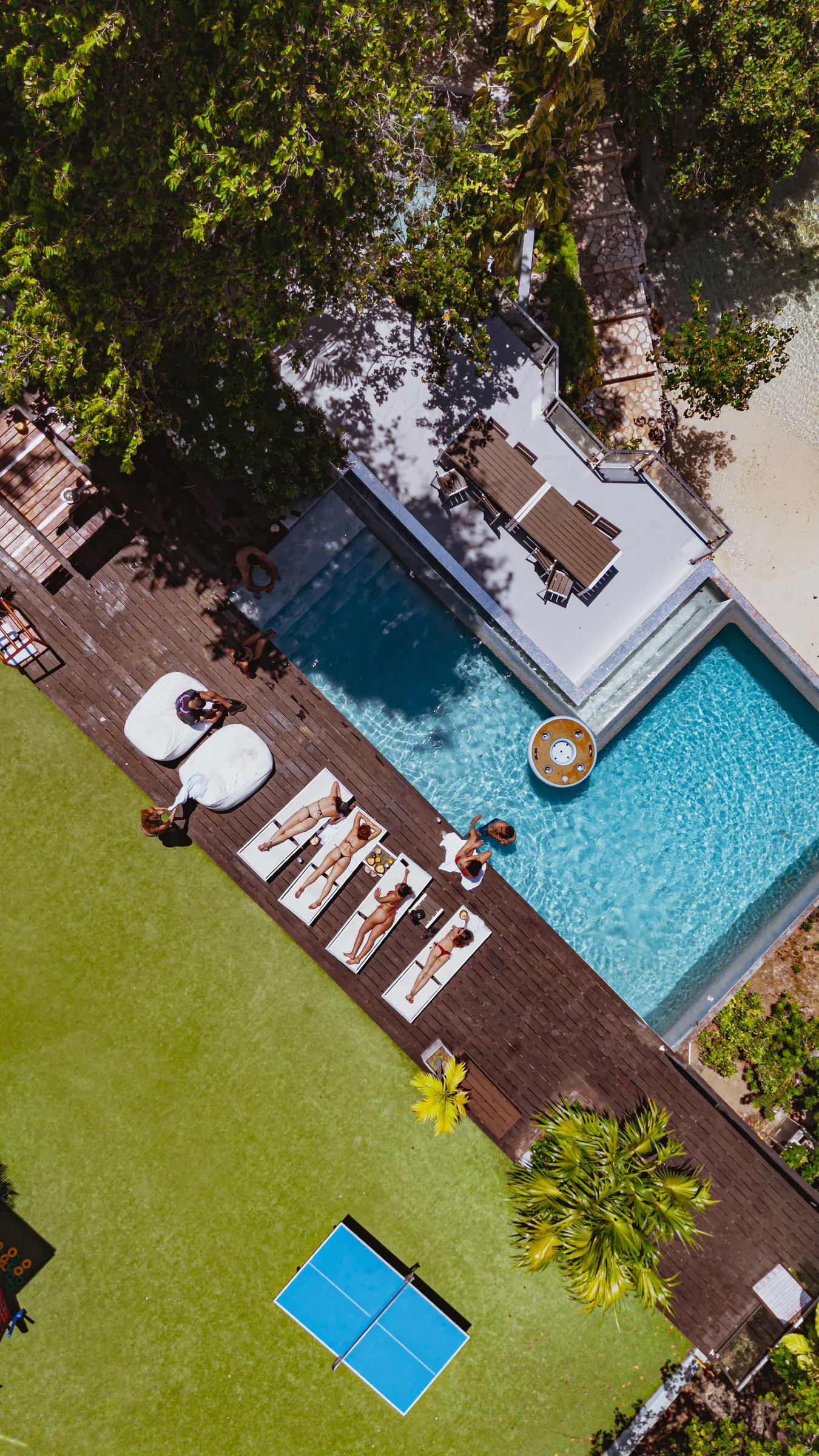 Straight down over a timber deck beside a pool, sunloungers and two white daybeds along one edge, a floating lounger on the water, lawn and a table tennis table beyond.