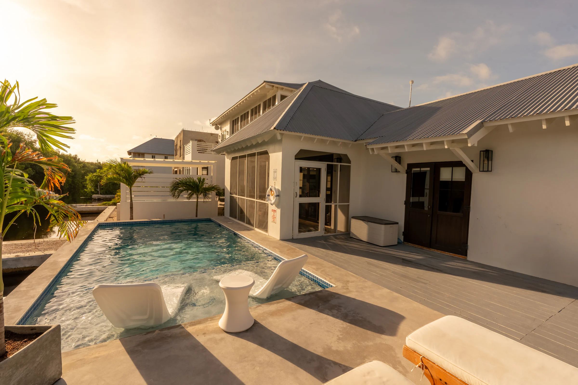 A rectangular pool with two submerged loungers, running up to a white villa with a grey metal roof and a screened porch, late afternoon sun behind the palms.