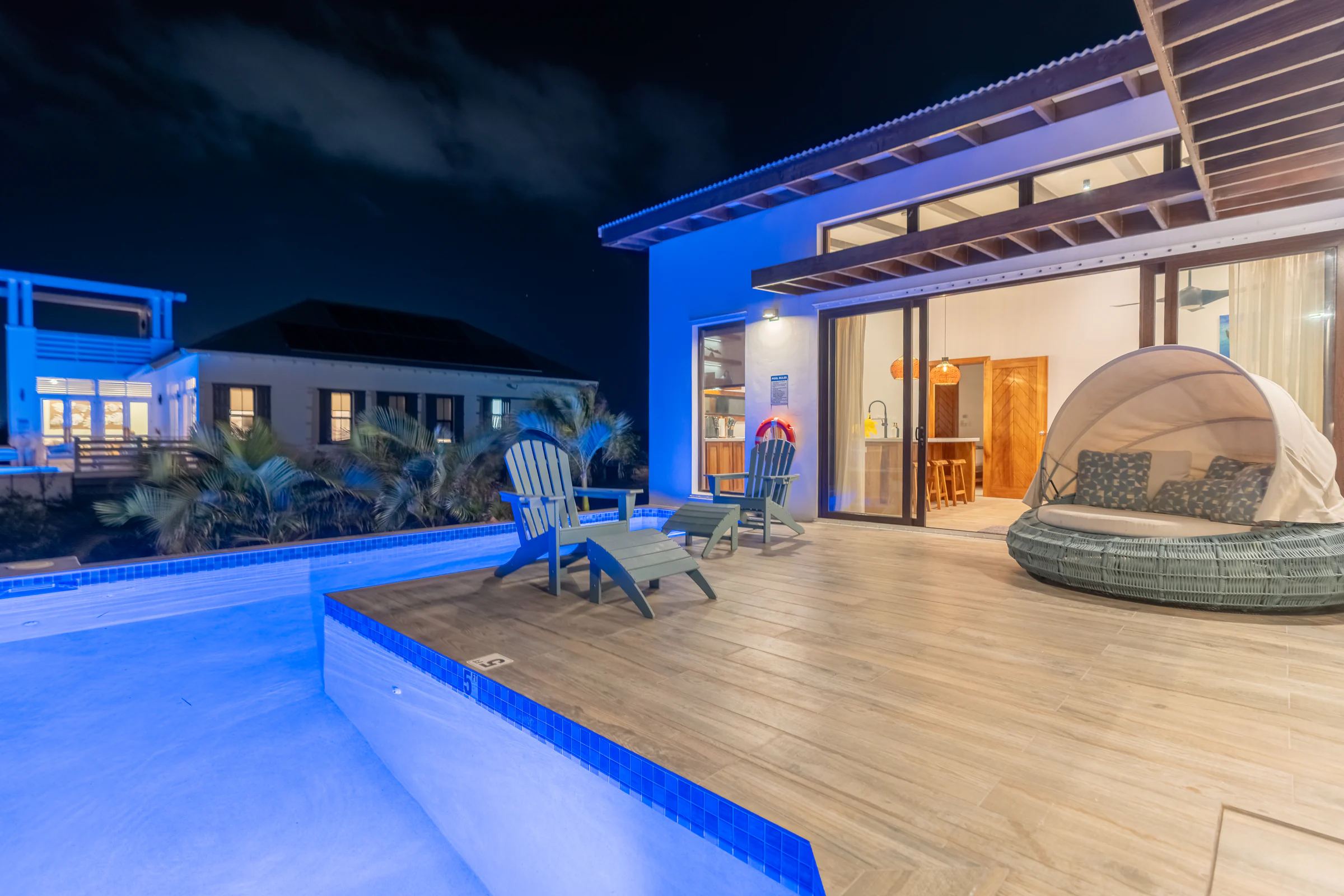 A pool deck after dark, the villa wall washed blue by the pool lights, Adirondack chairs and a canopied daybed on the timber deck, the kitchen lit warm behind open sliding doors.