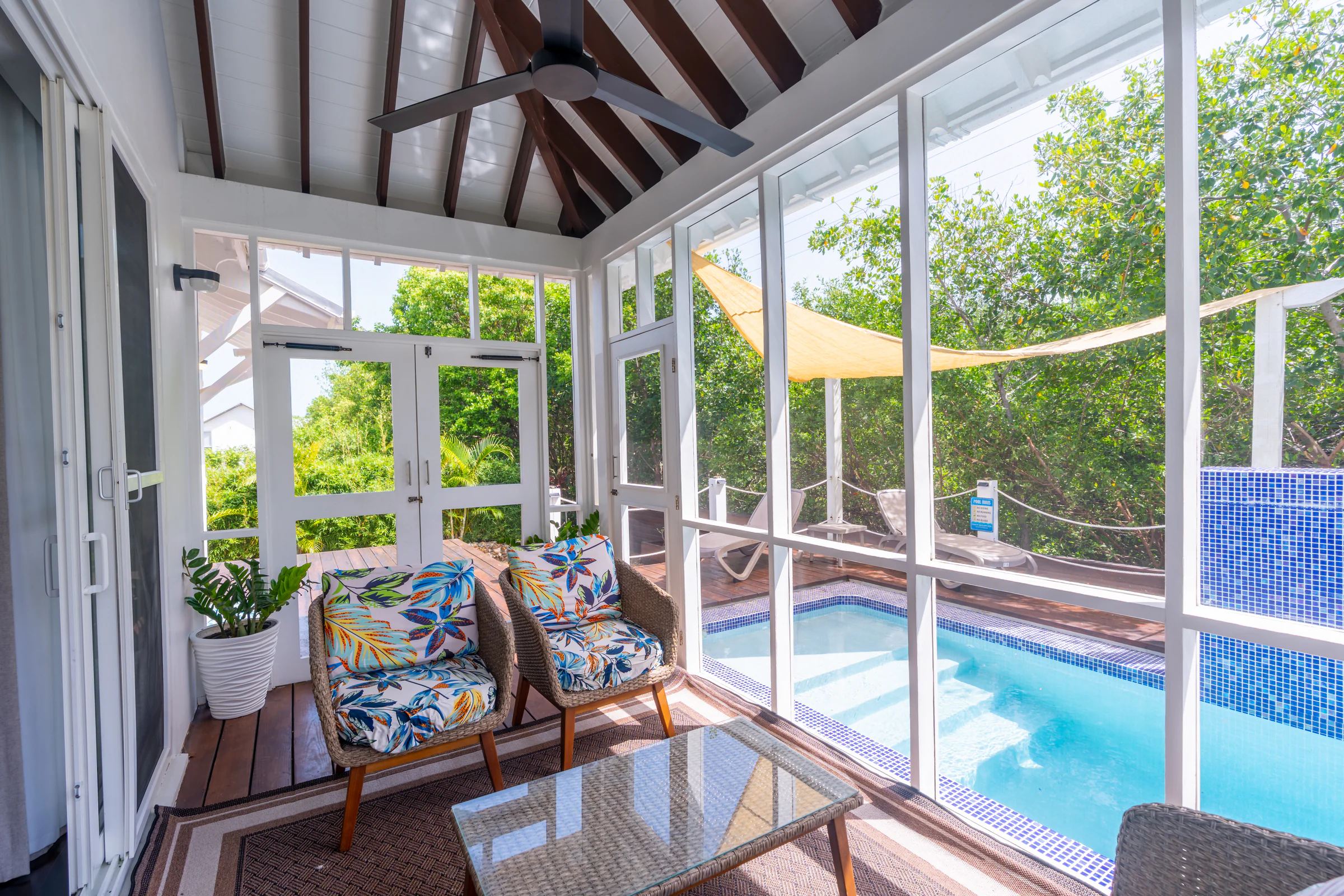 A screened porch with wicker chairs in tropical-print cushions and a ceiling fan, looking out through the screen to the pool and a shade sail under the trees.