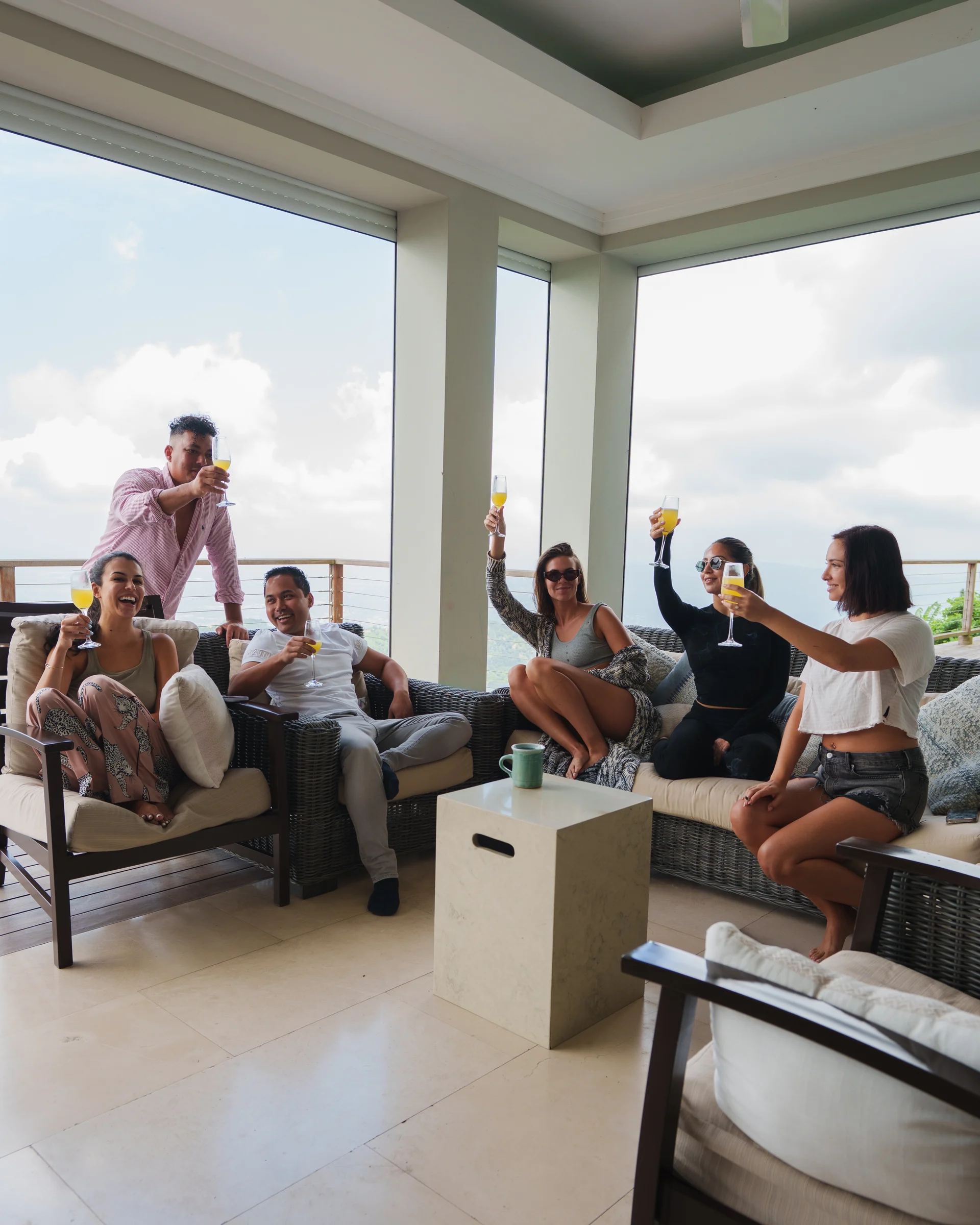 Five guests on a covered terrace raising mimosas over wicker sofas, floor-to-ceiling glass behind them open to cloud and hillside.