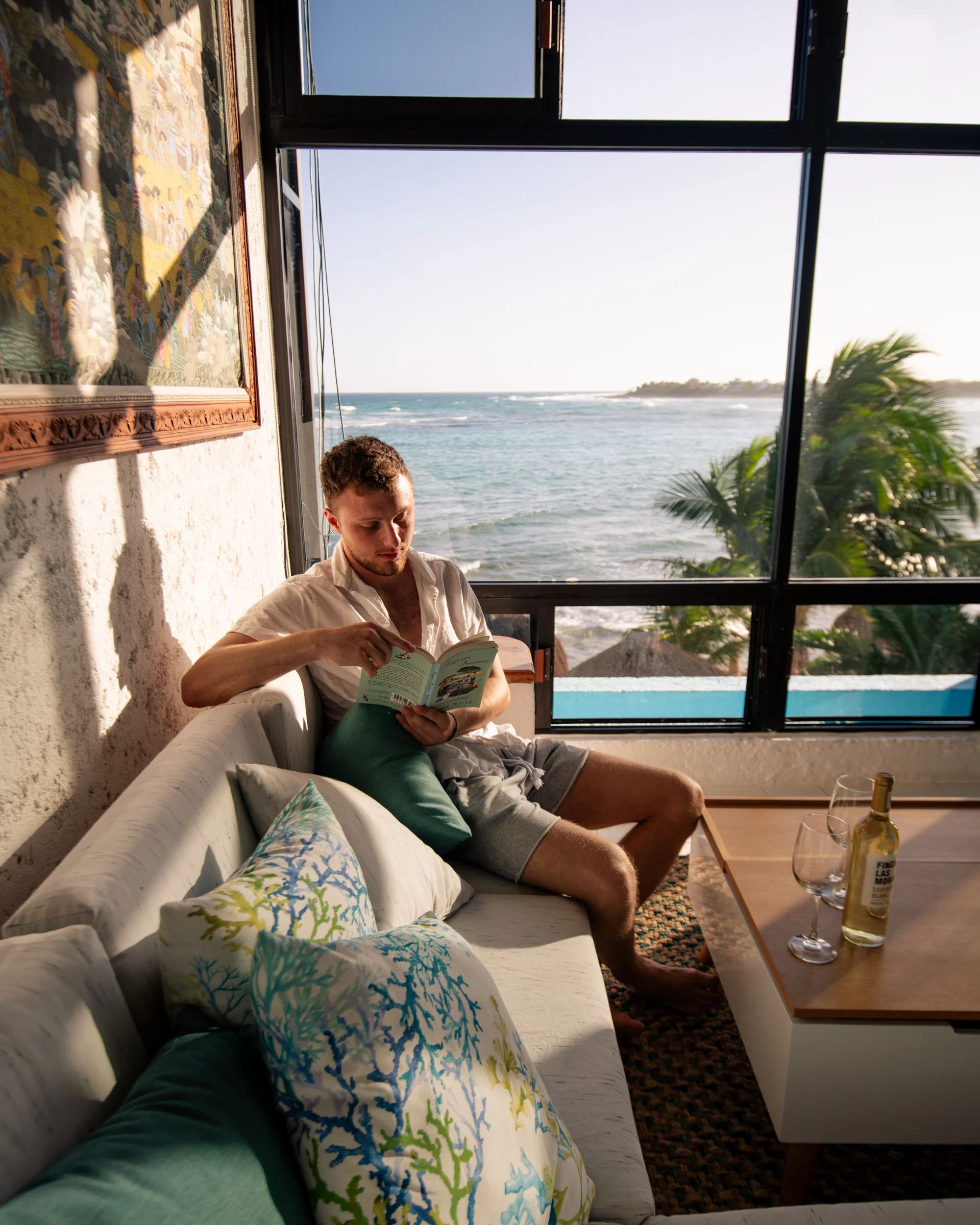 A guest reading on a built-in window seat in coral-print and teal cushions, the glass wall beside him open to a pool, palms, and the surf beyond, a bottle of white wine and two glasses on the low table.